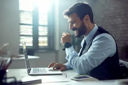 Homem de colete sorrindo enquanto usa notebook em mesa de escritório corporativo