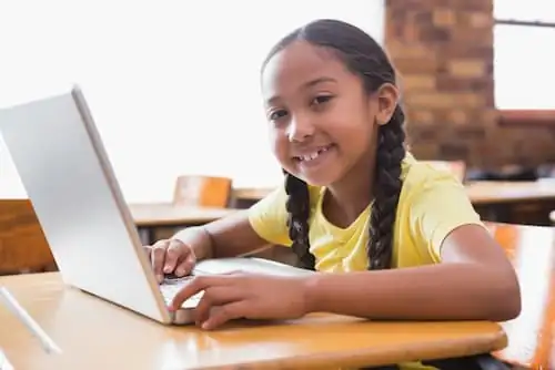 Menina sorrindo ao usar notebook na carteira escolar, ensino de computação na escola