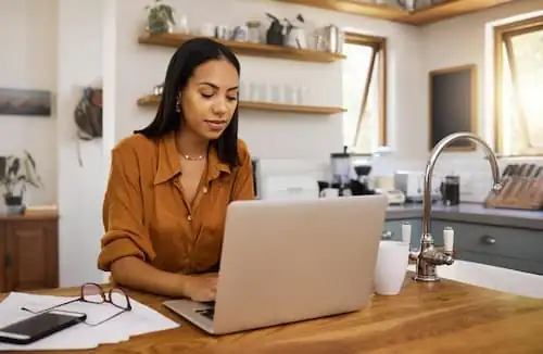 Mulher trabalhando em notebook na cozinha de casa, equipe em regime híbrido