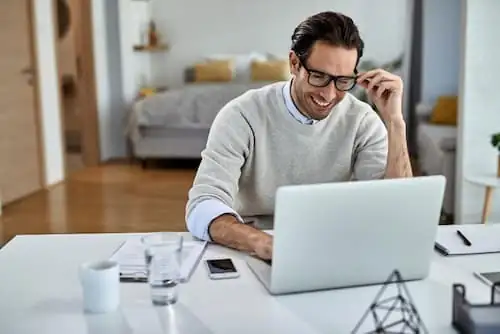 Homem sorrindo enquanto usa notebook em home office, trabalho híbrido